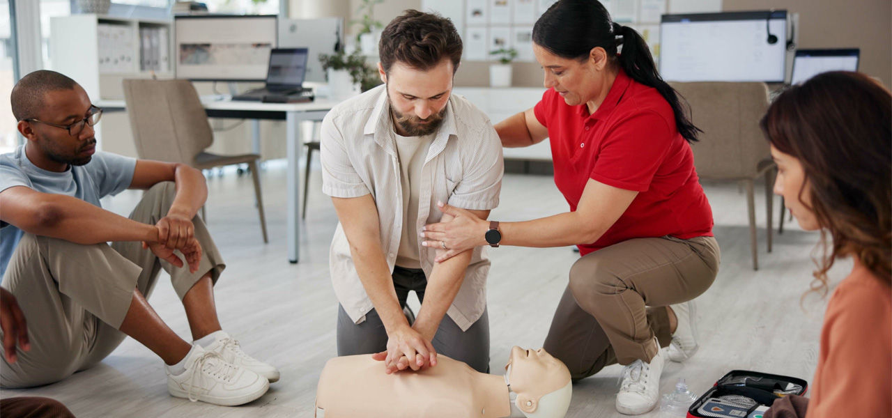 man practices CPR on a dummy in front of a CPR class while instructor coaches proper form