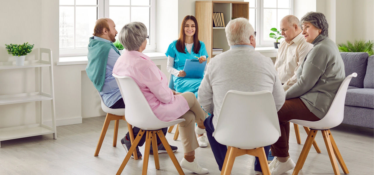 5 elderly people sit in chairs in a circle speaking with a healthcare provider in scrubs