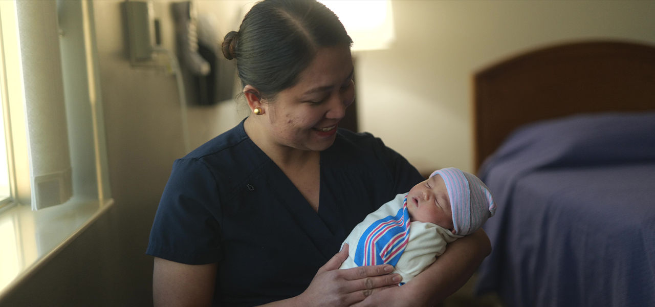 A nurse in dark scrubs gently holds a newborn baby wrapped in a striped hospital blanket.